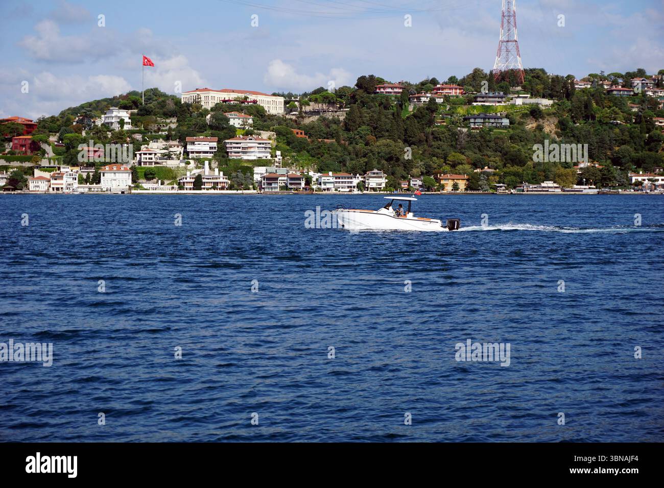 Istanbul, Türkei. Die größte Stadt in der Türkei und eine der wenigen transkontinentalen Städte der Welt, die sich über Europa und Asien auf der Bosporus-Straße erstreckt. Ein kleines weißes Motorboot mit rot-weiß gestreifter Markise, das auf einem tiefblauen Gewässer segelt. Das Boot bewegt sich in Richtung der rechten Seite des Bildes und hinterlässt eine weiße Spur. Im Hintergrund ist ein Hügel mit üppigen grünen Bäumen und Häusern zu sehen, mit einer roten Flagge, die auf einem der Gebäude thront. Ein hoher, weißer Turm mit einer rot-weiß gestreiften Spitze ist ebenfalls auf dem Hügel zu sehen. Der Himmel ist ein klares Blau mit ein paar verstreut Stockfoto
