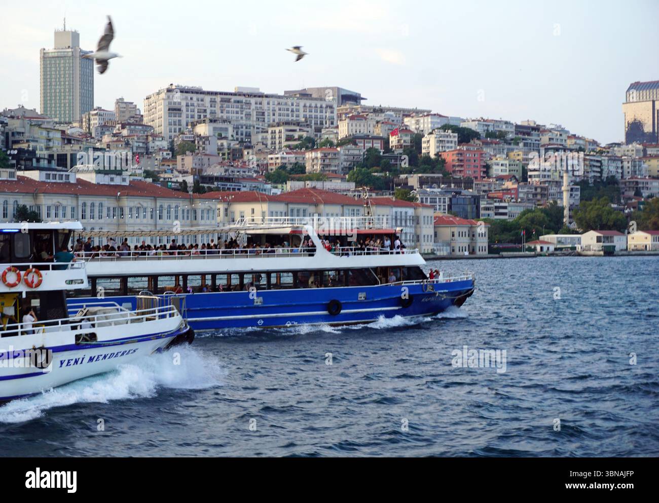 Istanbul, Türkei. Die größte Stadt in der Türkei und eine der wenigen transkontinentalen Städte der Welt, die sich über Europa und Asien erstreckt und über die Bosporus-Straße hinweg Eine pulsierende Stadtlandschaft vom Wasser aus betrachtet. Zwei Boote, ein blaues und ein weißes, sind im Vordergrund sichtbar, wobei das blaue Boot wie eine Fähre oder ein Passagierboot aussieht. Das weiße Boot hat einen rot-weiß gestreiften Abschnitt und den Namen „Yeni Mendes“ auf der Seite. Das Stadtbild im Hintergrund ist eine Mischung aus weißen und beigen Gebäuden mit roten Dächern, eingebettet auf einem Hügel. Das Wasser ist dunkelblau und der Himmel ist hellblau mit einem Stockfoto