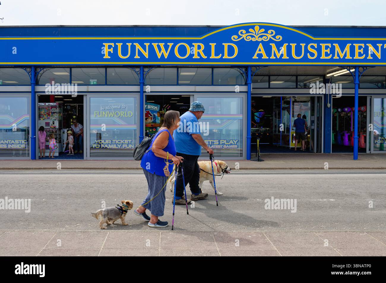 Ein fettleibiges älteres Paar mit Wanderstöcken und zwei Hunden auf der Leitung, die an der Funworld-Spielhalle an der Swanage-Küste Dorset England, Großbritannien, vorbeifahren Stockfoto