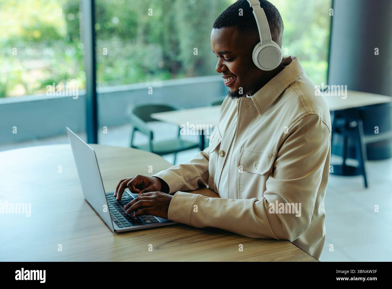 Ein lächelnder Geschäftsmann arbeitet ferngesteuert und verkörpert den Lebensstil digitaler Nomaden. Er trägt Kopfhörer und Typen auf seinem Laptop, sitzt in einem modernen Stil Stockfoto