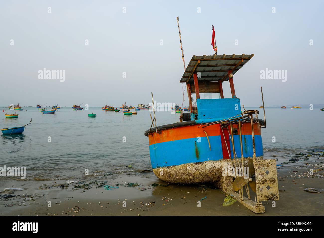Ein lebhaftes Fischerboot ruht auf dem Sand in Mui ne, Vietnam, während andere Boote auf dem ruhigen Meer am frühen Morgen schwimmen Stockfoto