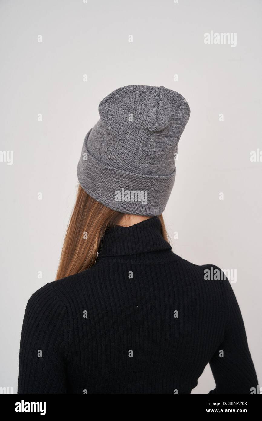 Graue Strickmütze, getragen von einer Person mit langen braunen Haaren, von hinten gesehen, in einem schwarzen Rippstrickpullover mit Rollkragen. Die Mütze ist lässig und Stockfoto