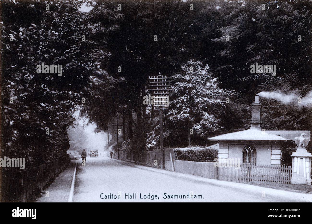 Eine ruhige, von Bäumen gesäumte Straße führt zur Carlton Hall Lodge in Saxmundham, deren Kamin sanft raucht. Ein rustikaler Zaun umgibt die Lodge, während ein Auto aus den frühen 1920er Jahren sich in der Ferne nähert – klein, aufrecht und erinnernd an eine neue Ära. Die Szene verbindet den edwardianischen Charme und moderne Verkehrsmittel. Von einer originalen, nicht zugeschriebenen Postkarte. Stockfoto