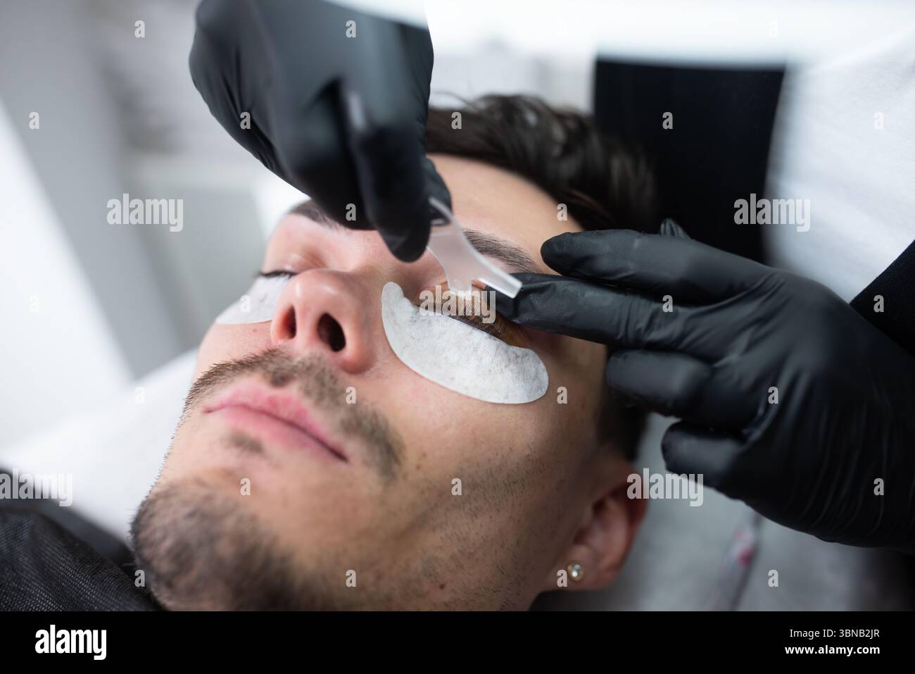 Männlicher Kunde wird in einem modernen Schönheitssalon einer Wimpernverlängerung unterzogen, wobei ein Kosmetiker sorgfältig Klebe- und Schutzpolster aufträgt Stockfoto