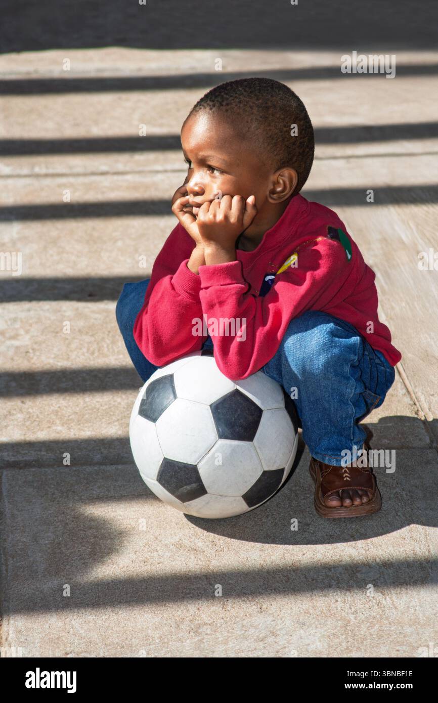 Trauriges afrikanisches Kind, ein Fußball zwischen seinen Beinen, Fäuste bedecken seinen Mund, Stockfoto