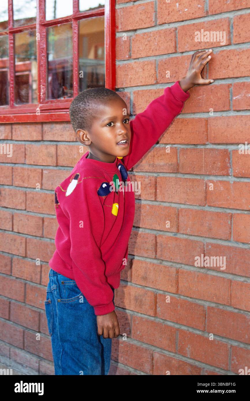 afrikanisches Kind, das draußen auf dem Schulhof eine Ziegelmauer hält, roter Pullover, Fenster hinten Stockfoto