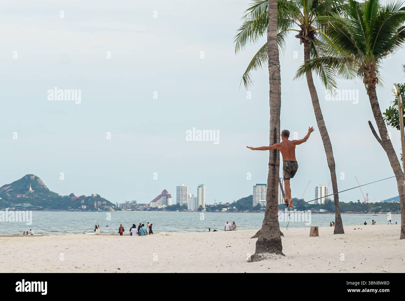 Ein Mann balanciert auf einem Drahtseil zwischen Palmen an einem Strand, mit einer Skyline von Hua hin in der Ferne, Thailand Stockfoto