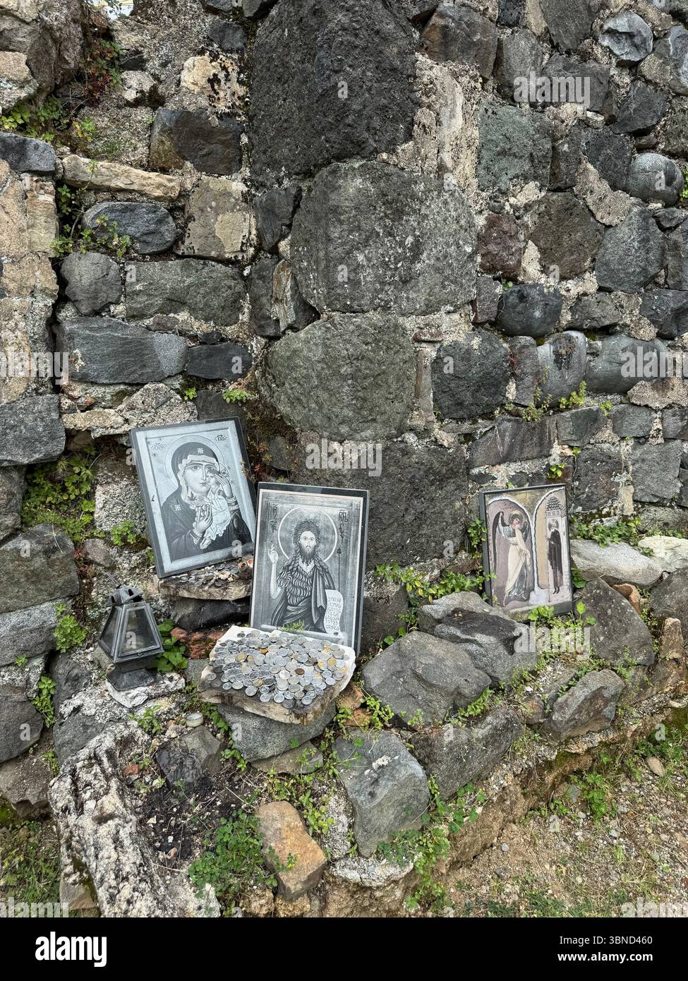 Dieses Foto zeigt drei religiöse Ikonen, einen Stapel gemischter Münzen und eine Laterne, die auf unebenen Steinen an einer alten Mauermauer angeordnet ist. Die Szene indiziert Stockfoto
