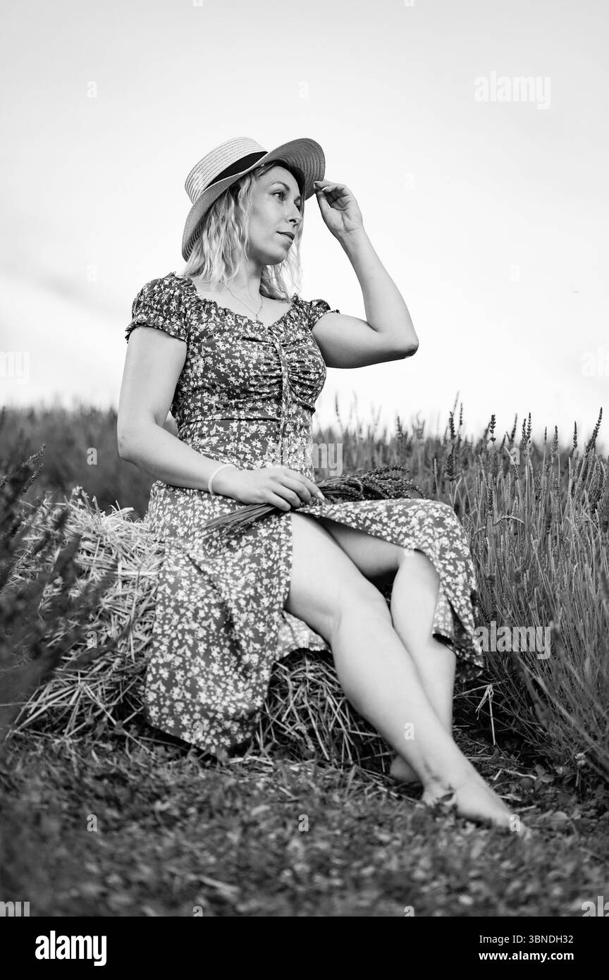 Ein heiteres Schwarz-weiß-Foto einer Frau mit Sommerkleid und Strohhut, die auf einem Heuballen sitzt und friedlich aussieht. Stockfoto