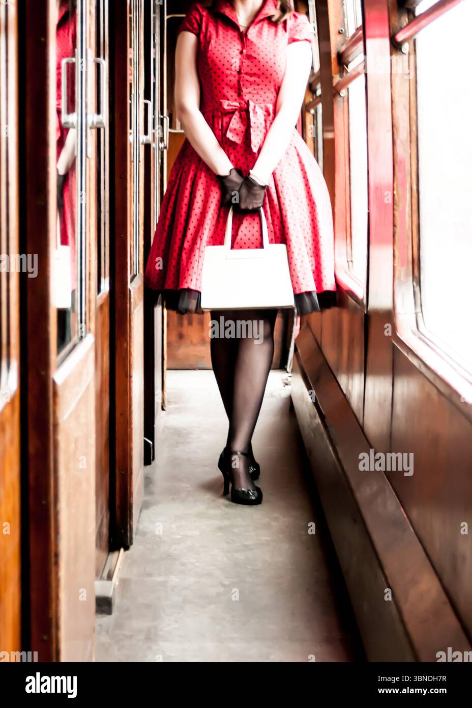 Eine Frau in einem roten Kleid mit Punktmuster, schwarzen Handschuhen und Absatzschuhen steht in einem Oldtimer-Zugwagen und hält eine weiße Handtasche. Stockfoto