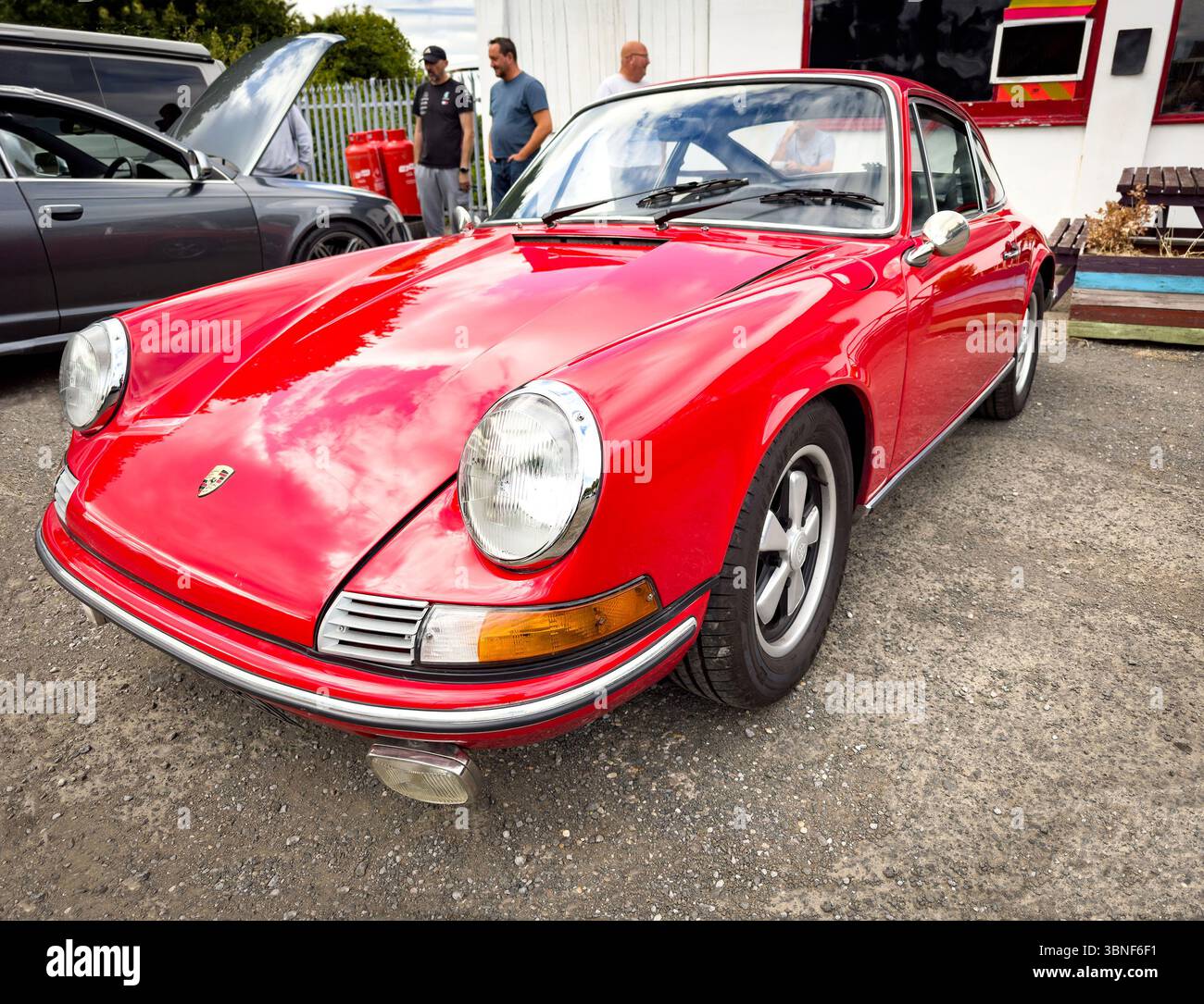 Klassischer, roter Porsche 911-Sportwagen mit elegantem Design und glänzender Oberfläche, der an einem bewölkten Tag auf Schotter neben anderen Fahrzeugen geparkt ist Stockfoto