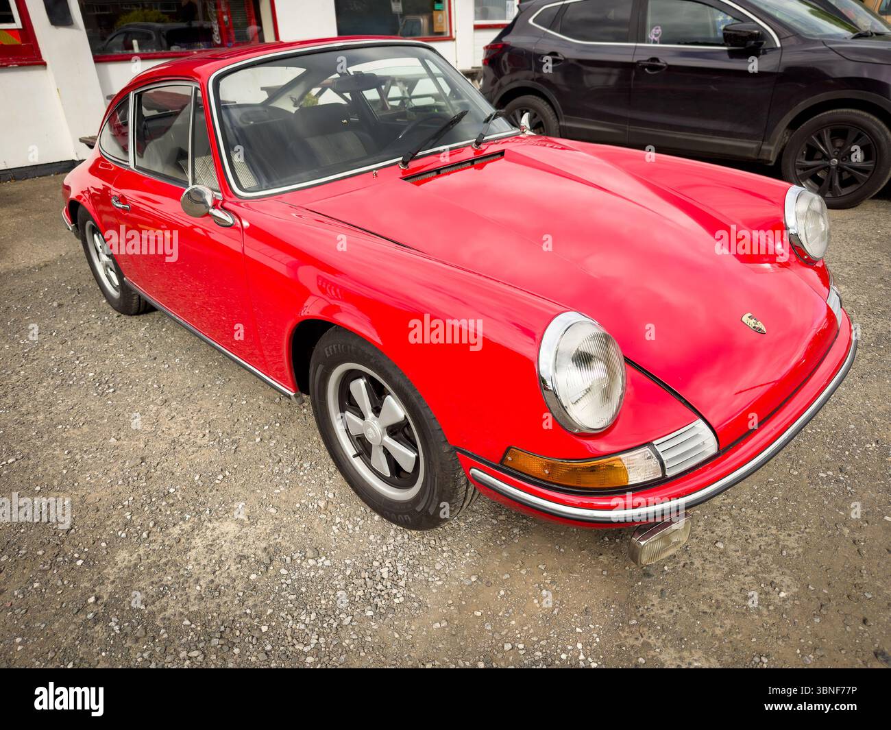 Klassischer, roter Porsche 911-Sportwagen mit elegantem Design und glänzender Oberfläche, der an einem bewölkten Tag auf Schotter neben anderen Fahrzeugen geparkt ist Stockfoto