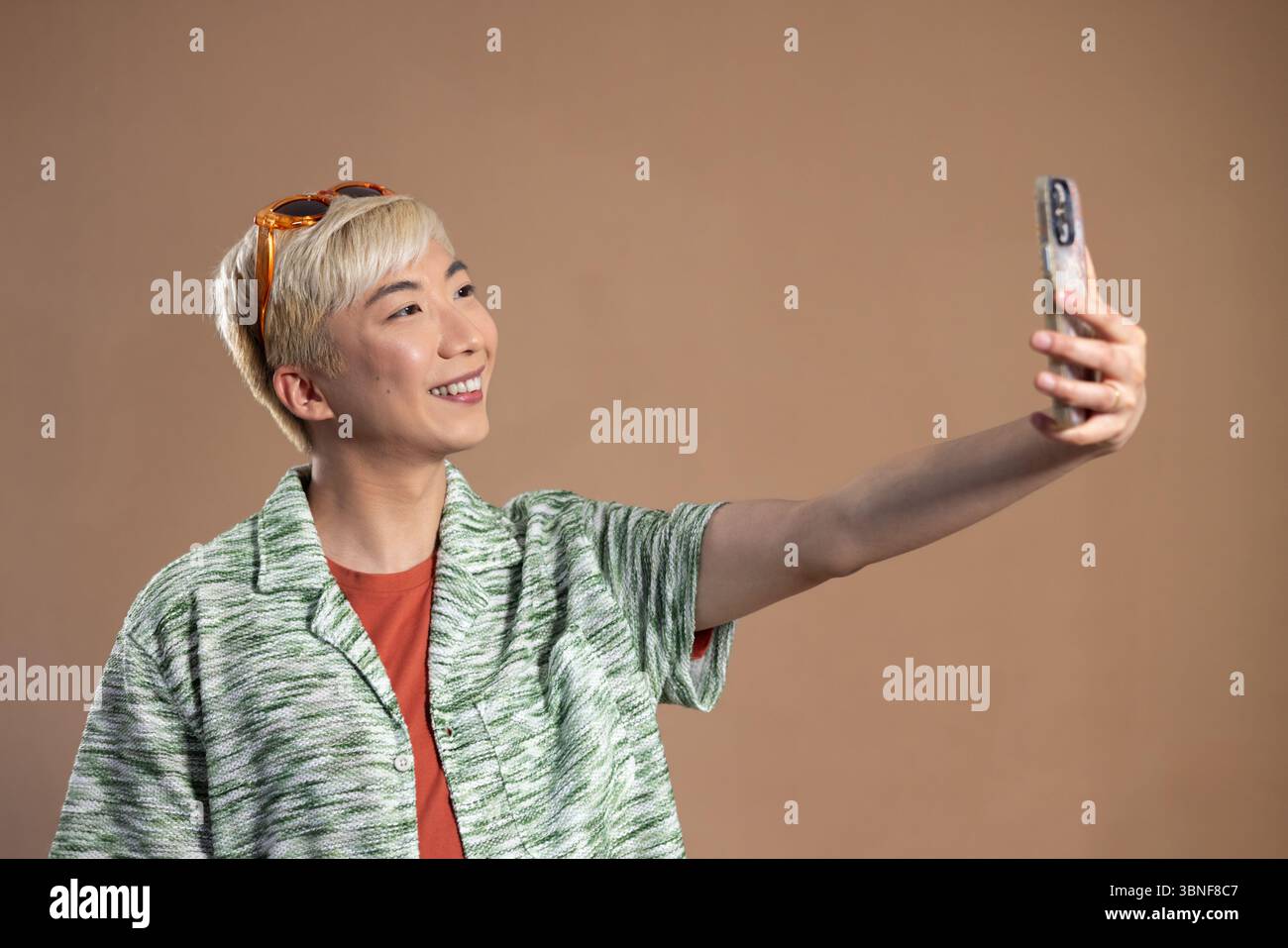 Studio-Aufnahme eines jungen erwachsenen Mannes, der für ein Selfie im Studio lächelt. Er hat gebleichte Haare und trägt ein gestricktes Hemd, steht gegen einen neutralen Stockfoto