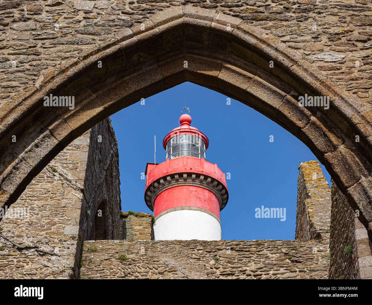 Rot-weißer Leuchtturm durch den Steinbogen der alten Abteiruinen Saint-Mathieu in der Bretagne, Frankreich, an einem sonnigen Nachmittag. Stockfoto