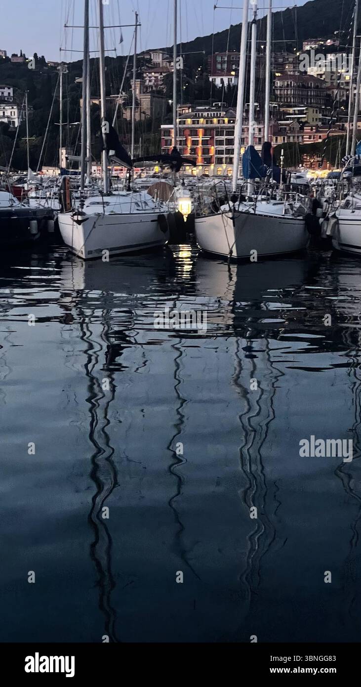 Segelboote schweben in der Abenddämmerung sanft in einem ruhigen Hafen und reflektieren das sanfte Licht von nahe gelegenen Gebäuden. Ruhiges Wasser spiegelt die Masten und schafft ein ruhiges e Stockfoto