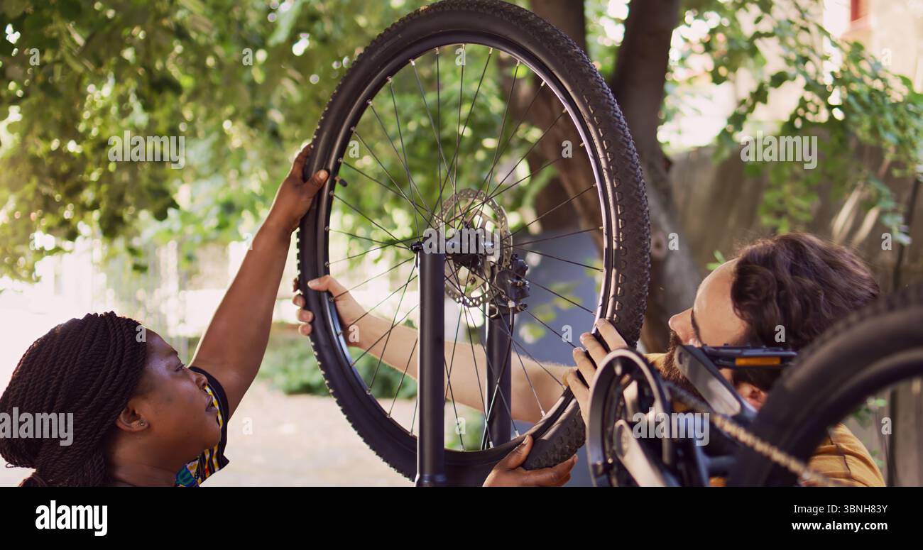 Die afroamerikanische Freundin trägt ein Rad, um sie mit ihrem kaukasischen Freund wieder an der fahrradkarosserie zu befestigen. Sportliebendes multiethnisches Paar, das den demontierten Fahrradreifen draußen sorgfältig sichert. Stockfoto