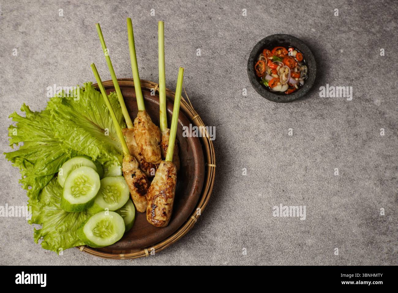 Traditioneller Satay lilit aus Bali, serviert mit Gurke, Salat und Sambal Matah in einer Steinschale. Aufgenommen auf einem strukturierten grauen Hintergrund. Stockfoto