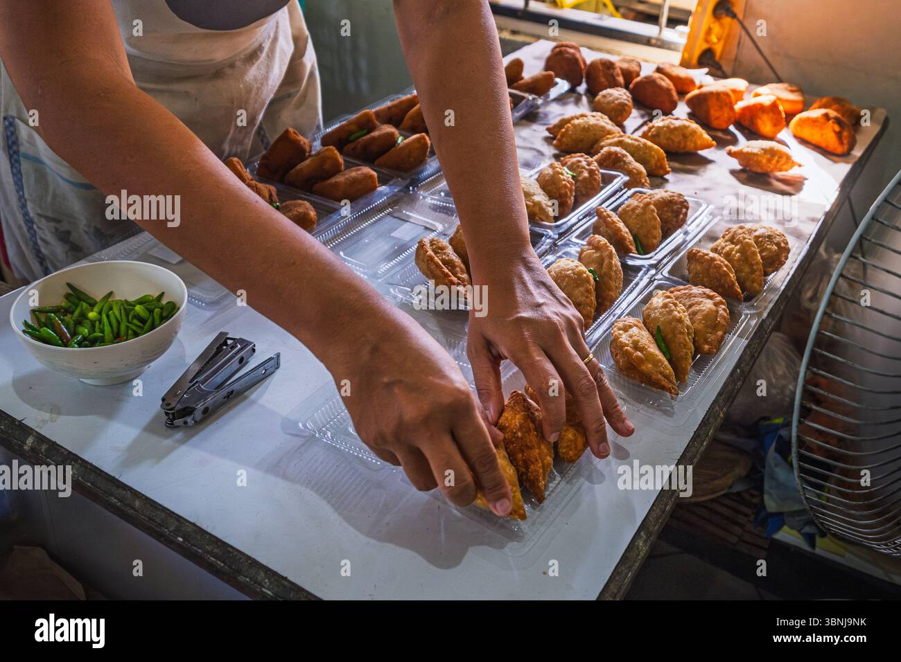 Nahaufnahme der Hände einer Frau, die traditionelle indonesische frittierte Snacks, Pastell und Rillen sorgfältig in Plastikbehältern als Teil einer kleinen Ho arrangieren Stockfoto