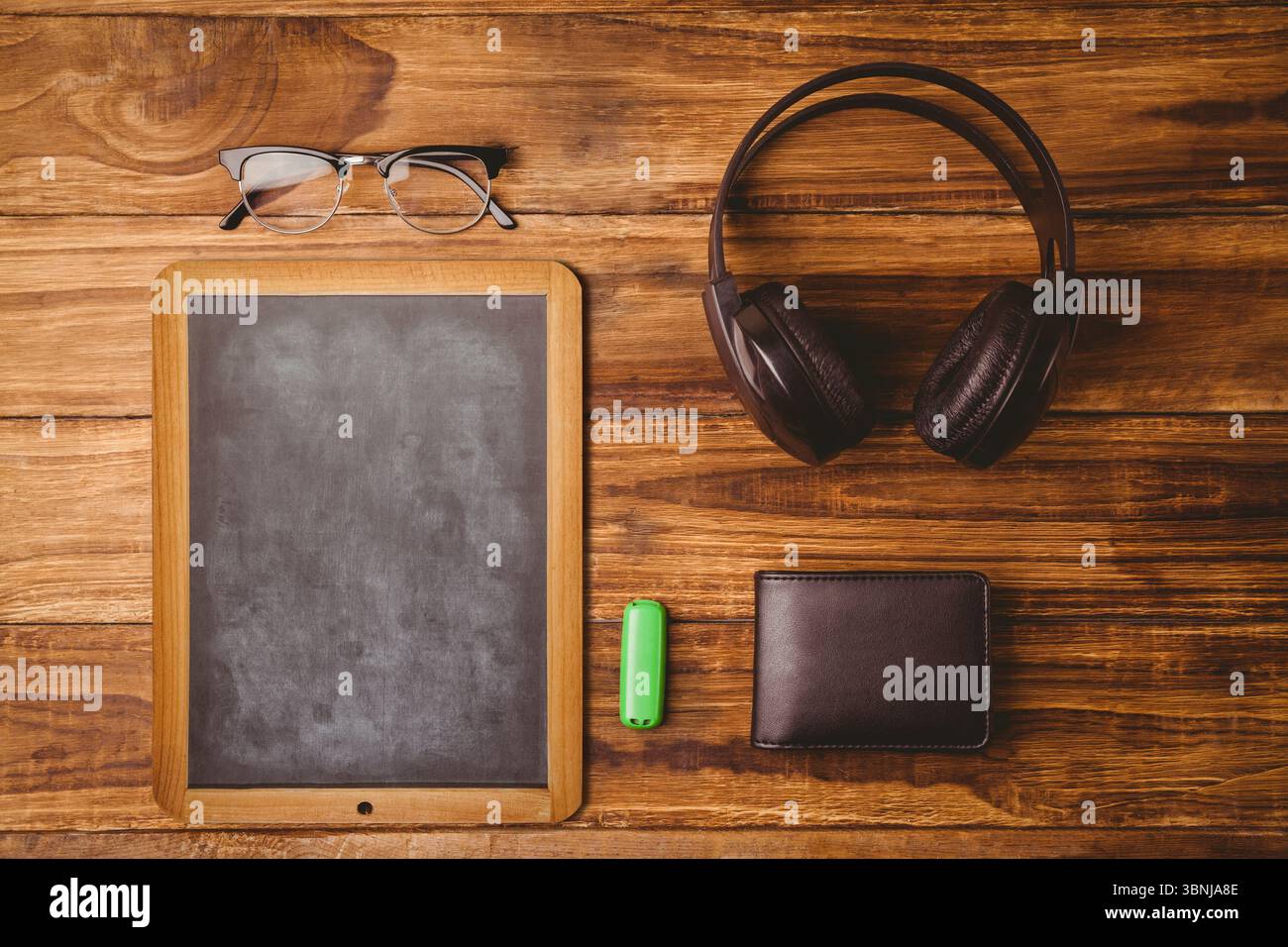 Die kleine Tafel sitzt auf einem Holzschreibtisch in flachem Design und zeigt Brillen, Kopfhörer, Geldbörse und USB Stockfoto