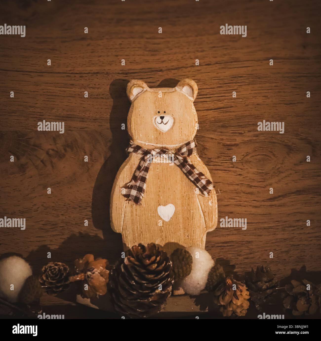 Ein gemütliches, vertikales Stillleben mit rustikaler Holzbärendekoration mit kariertem Schal und weißem Herz. Tannenzapfen und unscharfe Elemente. Stockfoto