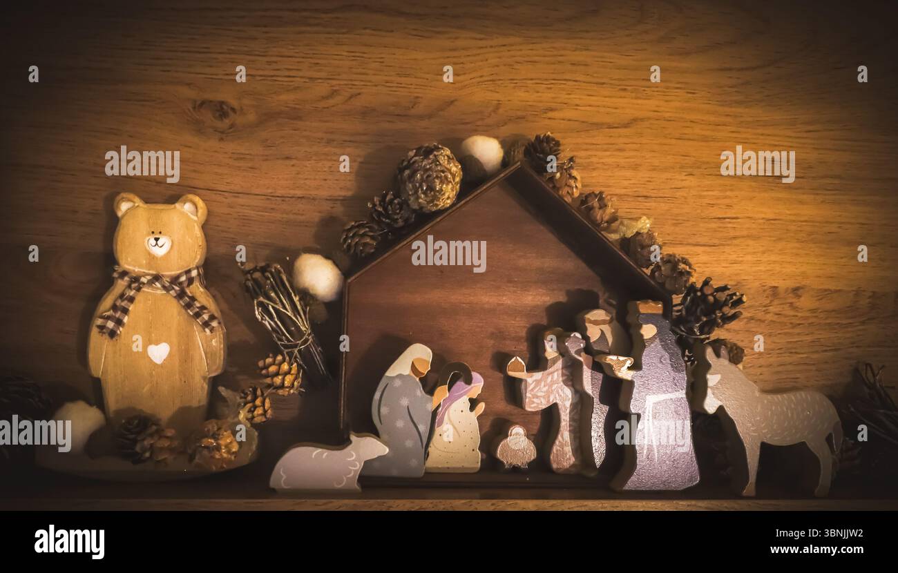Eine rustikale Weihnachtskrippe aus Holz wird auf einem Regal vor einer Holzwand ausgestellt, die von warmem Licht beleuchtet wird. Das Kunstwerk stellt die Heilige Familie dar Stockfoto