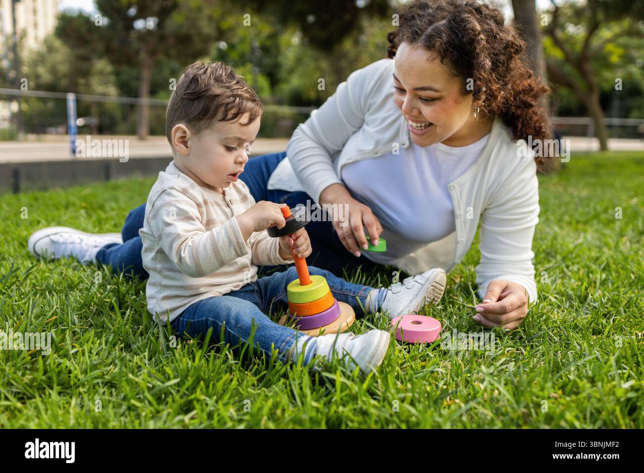 Eine Mutter und ihr Baby genießen einen sonnigen Tag im Park. Auf dem Gras teilen sie einen fröhlichen Moment, während das Baby bunte, aufgestapelte Spielzeuge erforscht Stockfoto