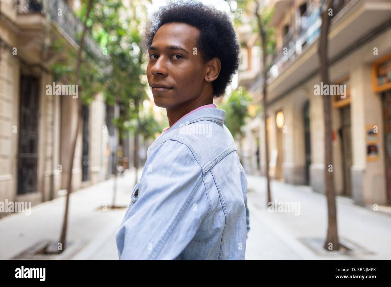 Eine nicht-binäre Person afroamerikanischer Herkunft geht selbstbewusst eine sonnenbeschienene Stadtstraße hinunter und trägt eine leichte Jeansjacke. Die Szene fängt Diversi ein Stockfoto