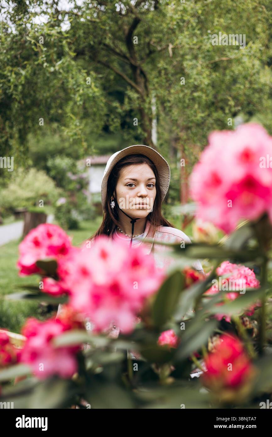 Eine Frau steht in einem üppigen Garten, umgeben von leuchtenden rosa Rhododendronblüten. Sie trägt einen Hut, der harmonisch mit dem ruhigen natürlichen Settin harmoniert Stockfoto