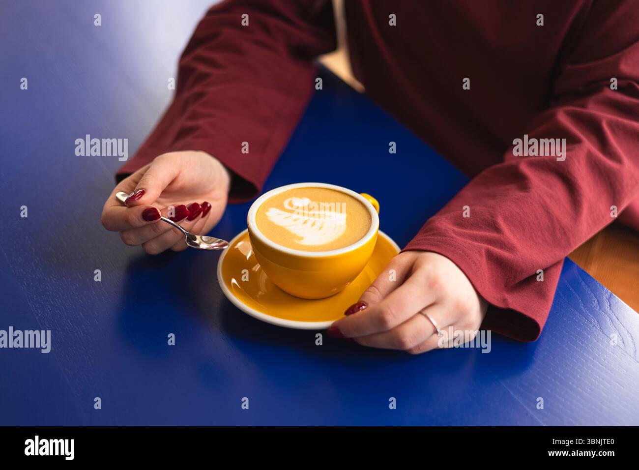 Eine Frau genießt ihren Kaffee in einem Café, in dem komplexe Schwanenlatte-Kunst in einer leuchtend gelben Tasse gezeigt wird. Die Szene wird vor einem markanten blauen Tisch inszeniert Stockfoto