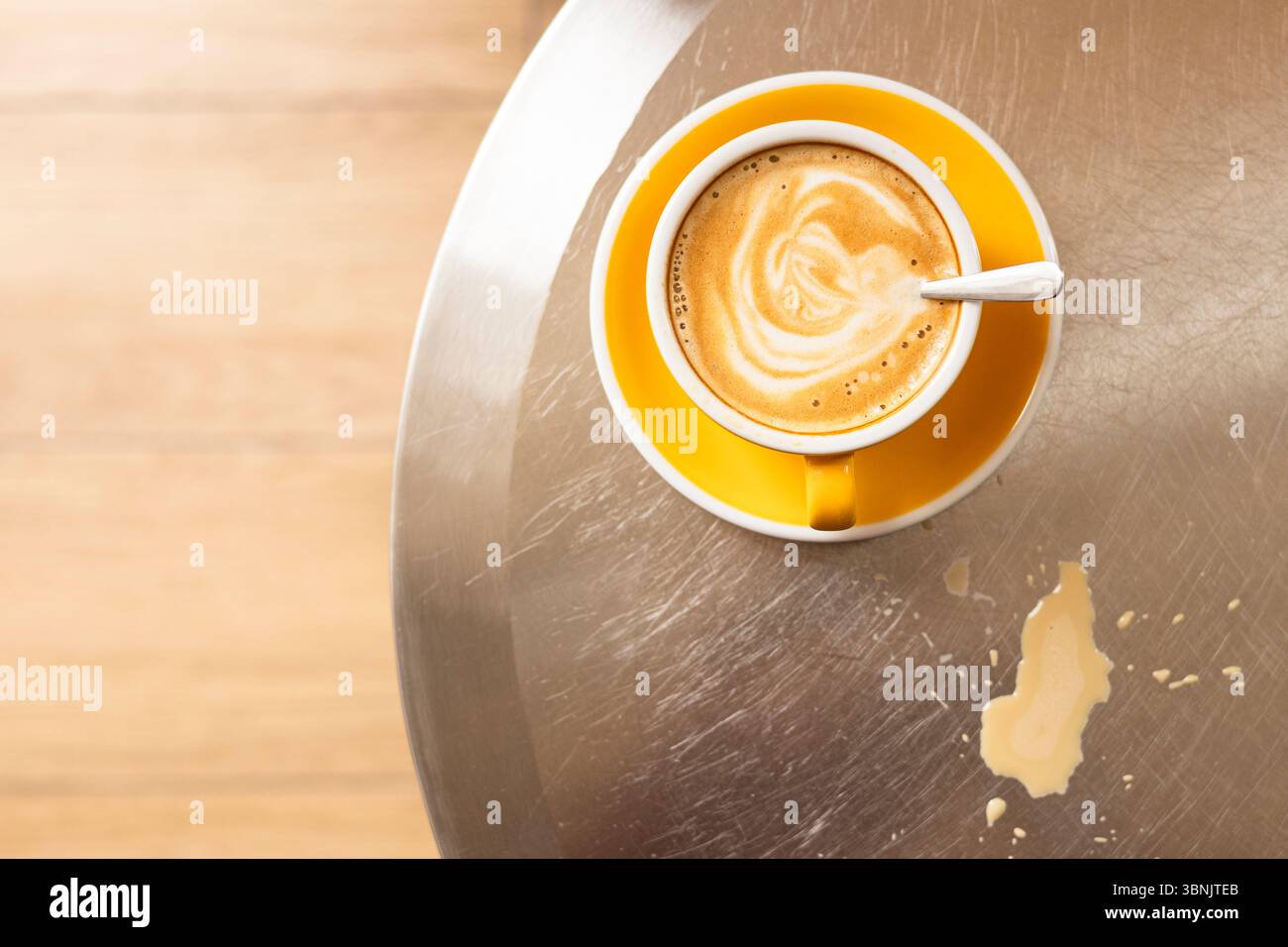 Eine leuchtend gelbe Tasse mit Kaffee sitzt auf einer reflektierenden Metallic-Oberfläche. Daneben trägt ein Kaffeefleck zum rustikalen Charme bei. Die Komposition exu Stockfoto