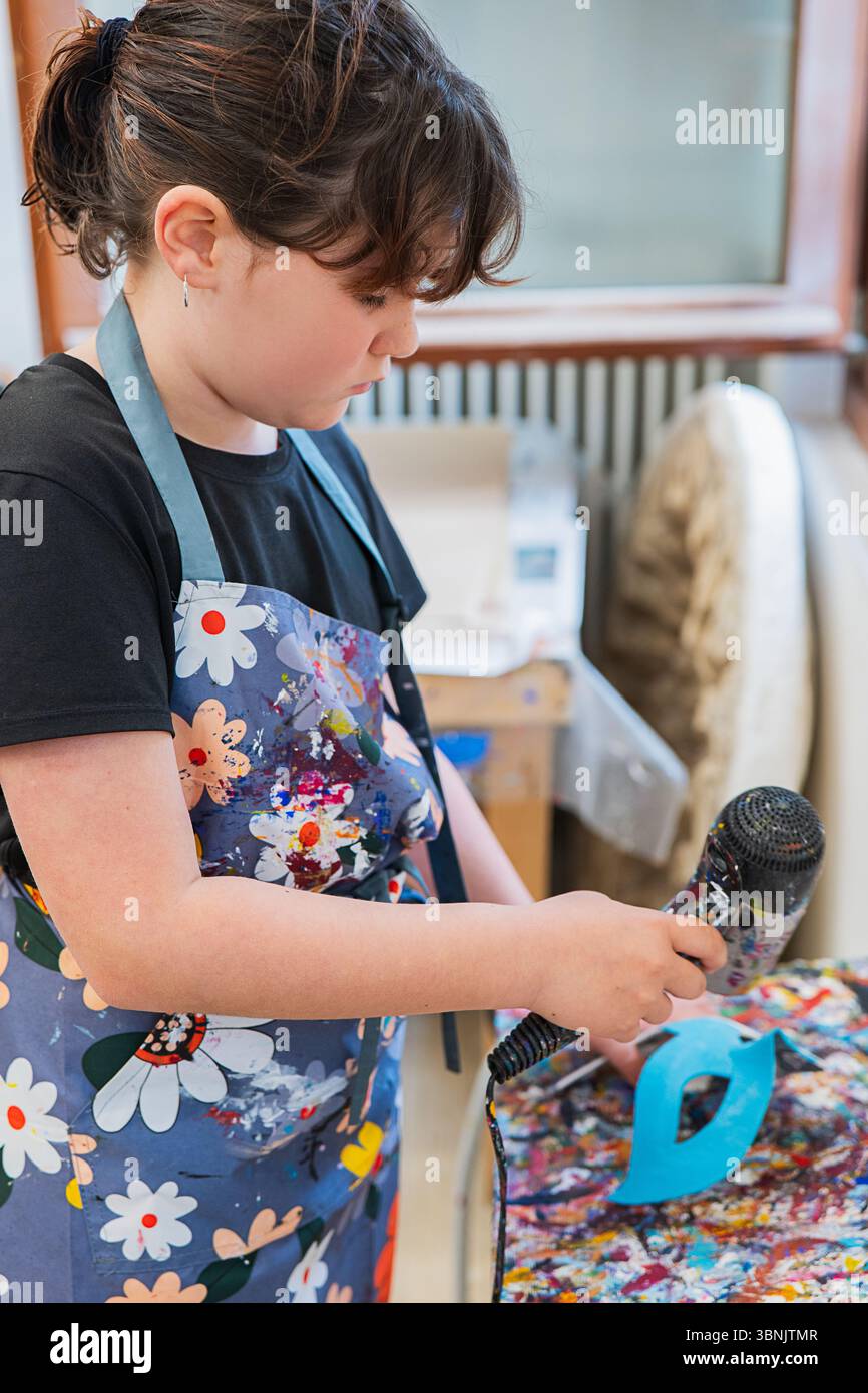 Ein Kind nimmt an einer lebhaften Maskenwerkstatt in Venedig Teil, die einen dekorierten Haarfön verwendet. Die florale Schürze verleiht diesem Kreativ einen Farbtupfer Stockfoto