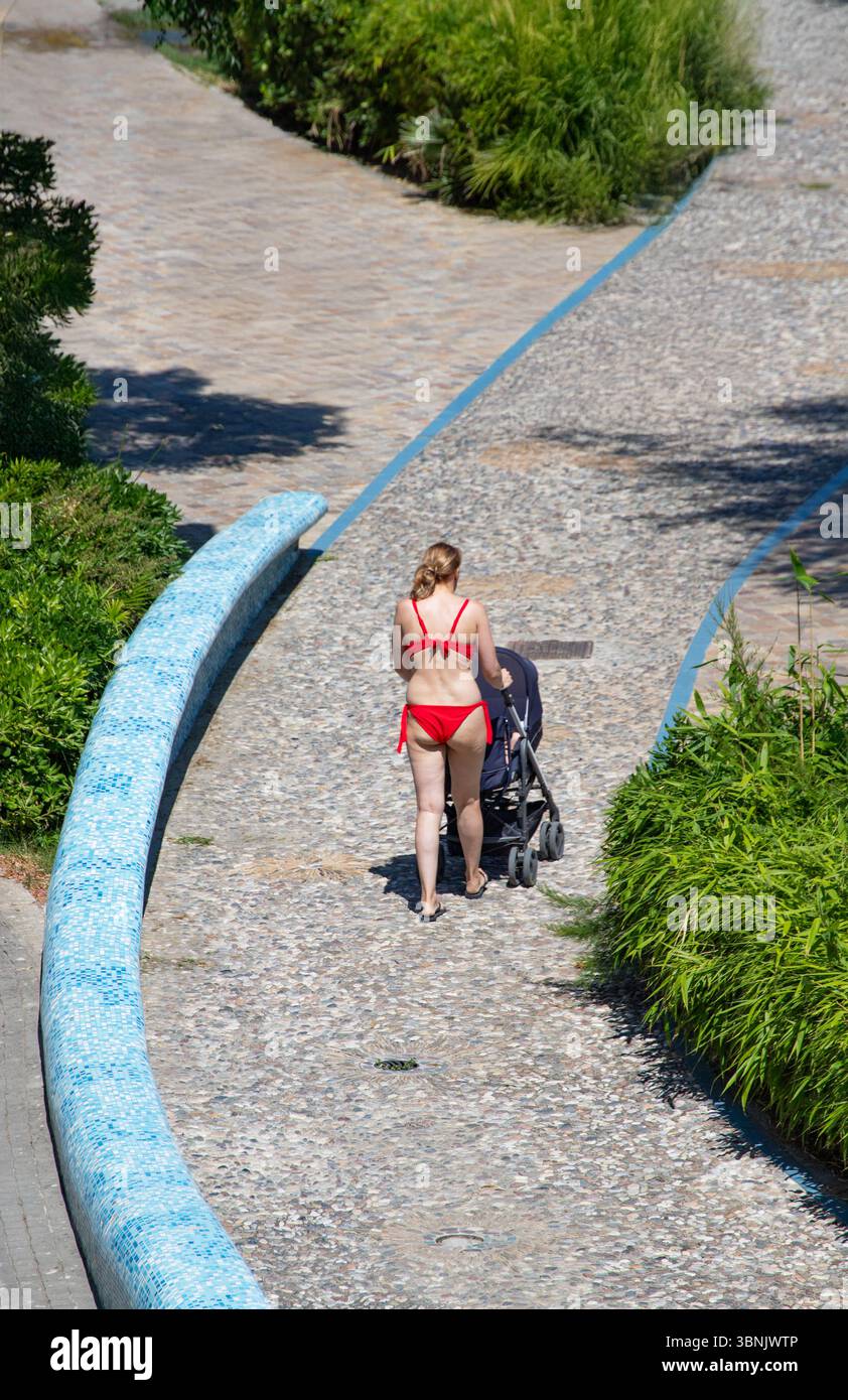 Frau schiebt Kinderwagen auf gepflasterten Weg und genießt sonnige Outdoor Walk grüne Pflanzen Sommertag. Stockfoto
