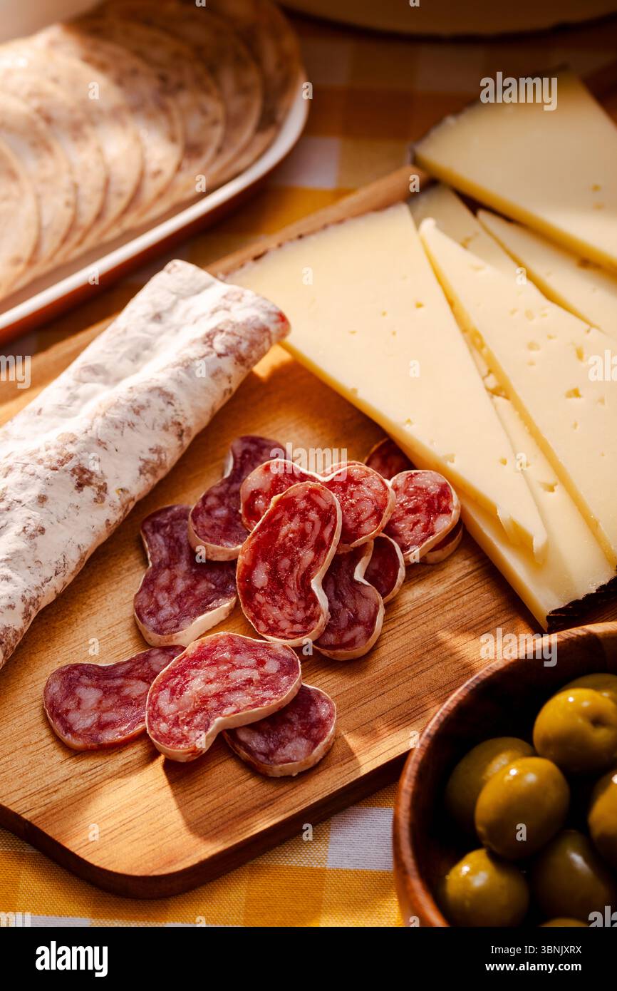 Eine rustikale Holzplatte mit katalanischem Fuet und gereiftem Käse, ergänzt durch eine Schüssel mit grünen Oliven. Perfekt für ein traditionelles Mittelmeer Stockfoto