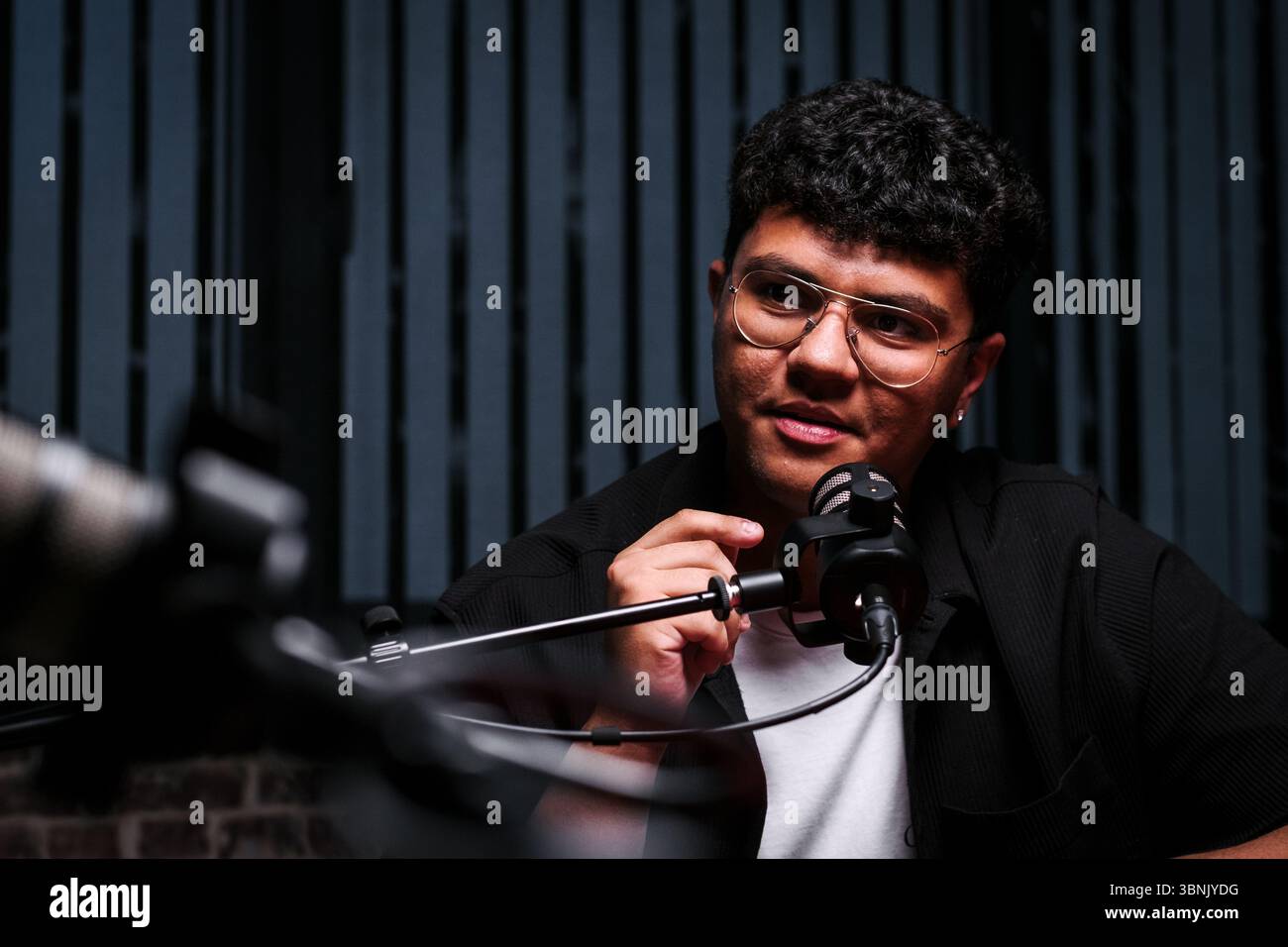 Ein Podcast-Host, der eine Brille trägt, spricht während einer Live-Aufnahme in ein Mikrofon. Das Setting verfügt über professionelle Audiogeräte, die noch besser sind Stockfoto