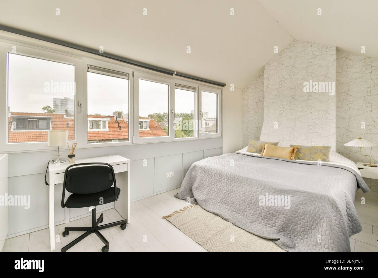 Helles und luftiges Schlafzimmer mit moderner Einrichtung, komfortablem Bett mit grauer Bettwäsche, weißem Schreibtisch mit schwarzem Stuhl und großen Fenstern Stockfoto