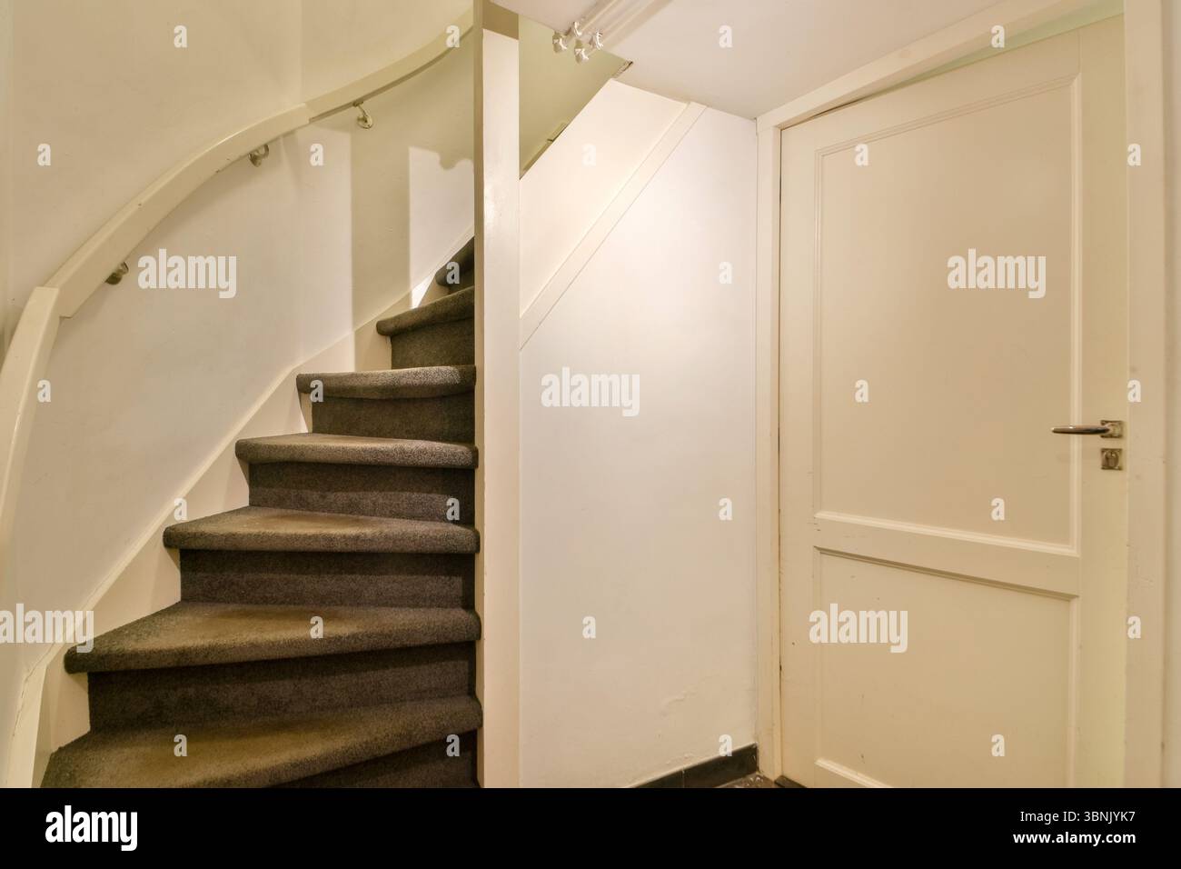 Eine gemütliche Inneneinrichtung mit einer geschwungenen Treppe mit Teppichboden neben einer geschlossenen weißen Tür. Das Ambiente strahlt eine schlichte und klare Ästhetik aus. Stockfoto