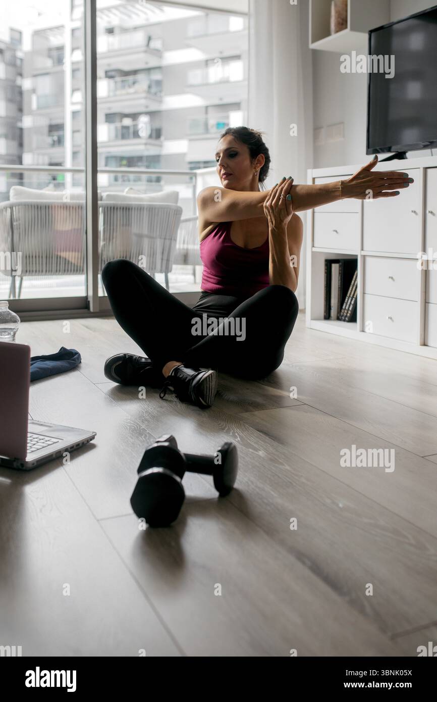 Eine Frau dehnt sich während des Trainings zu Hause, mit Hantel und Laptop in der Nähe, die moderne Fitnessroutinen und den Komfort des Heimtrainings hervorheben Stockfoto