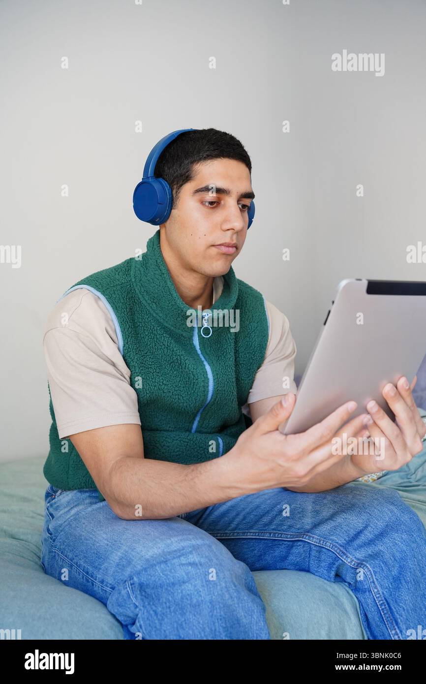 Ein junger arabischer Mann sitzt auf einem Bett, tief in sein Tablet verwickelt, während er blaue Kopfhörer trägt. Er ist lässig gekleidet, in seinen täglichen Rutt vertieft Stockfoto