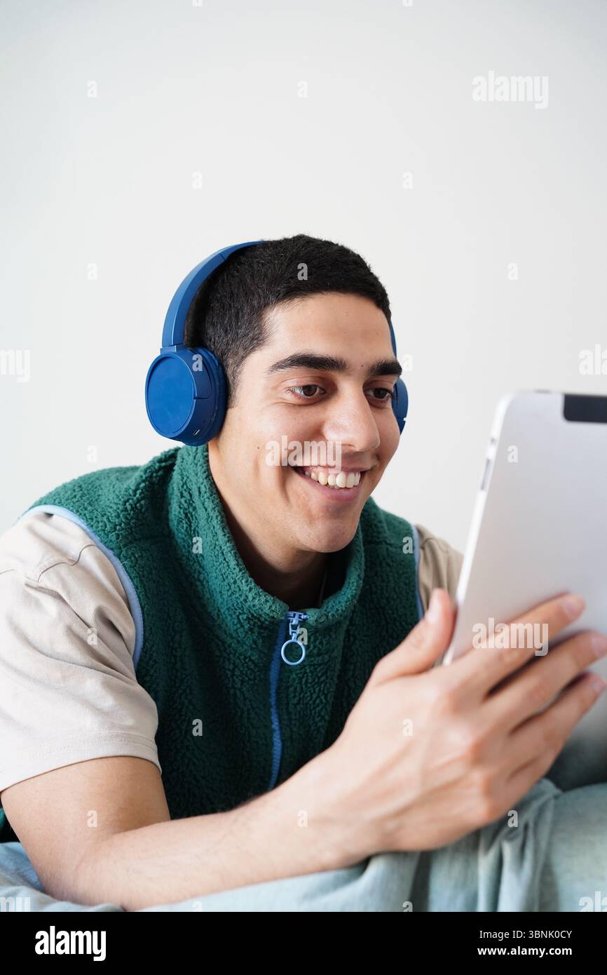 Junger arabischer Mann, der seine tägliche Routine genießt und Kopfhörer trägt, während er mit einem Tablet interagiert. Er ist entspannt und lächelnd, eingetaucht in ein digitales Erlebnis Stockfoto