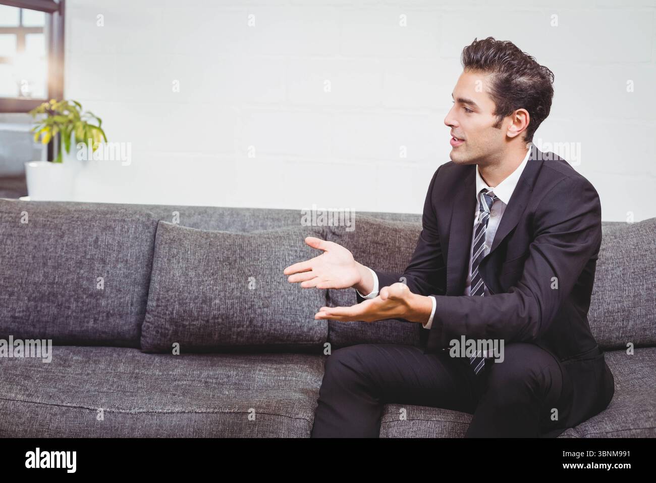 Graues Polstersofa, das an einer weißen Ziegelwand unter einem Holzfenster mit Topfpflanzen anliegt Stockfoto