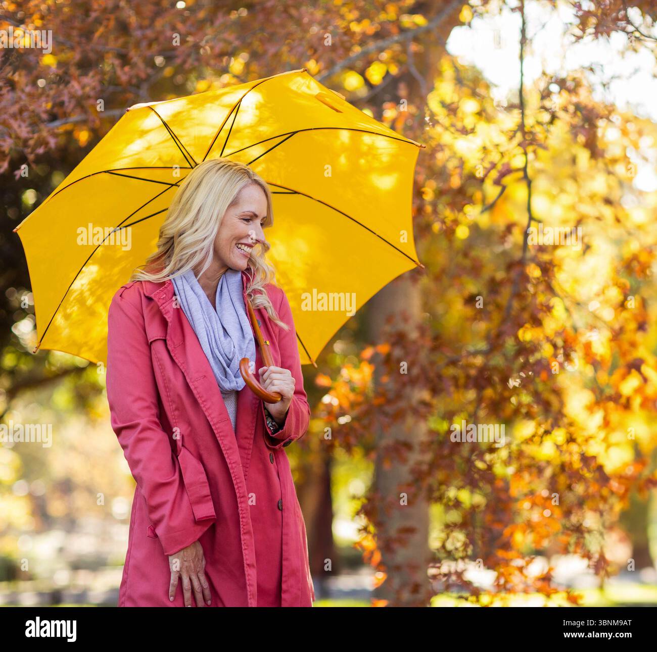 Lächelnde Frau in rotem Mantel, grauem Schal, gelber Schirm im Park zwischen goldenen Herbstblättern Stockfoto