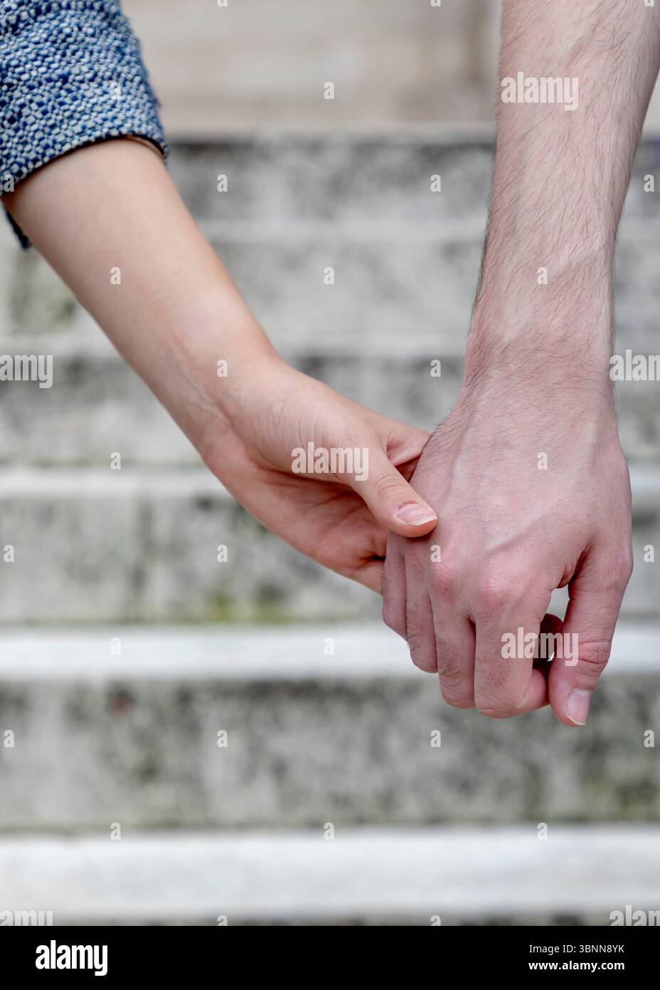 Eine Nahaufnahme von zwei Personen, die Hände halten, mit Fokus auf ihre Hände und Unterarme. Der Hintergrund ist verschwommen, scheint aber eine Treppe zu sein. Stockfoto