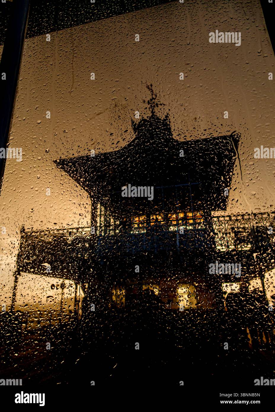 Die Silhouette eines Gebäudes ist durch ein regenbedecktes Fenster zu sehen. Das Gebäude hat ein einzigartiges architektonisches Design und der Regen erzeugt einen strukturierten Effekt Stockfoto