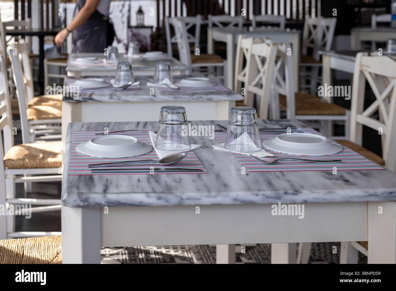 Leere Restauranttische mit weißen Marmorplatten sind ordentlich mit Tellern, umgedrehten Gläsern und Besteck besetzt und warten auf Gäste in einem hellen, einladenden Ambiente Stockfoto