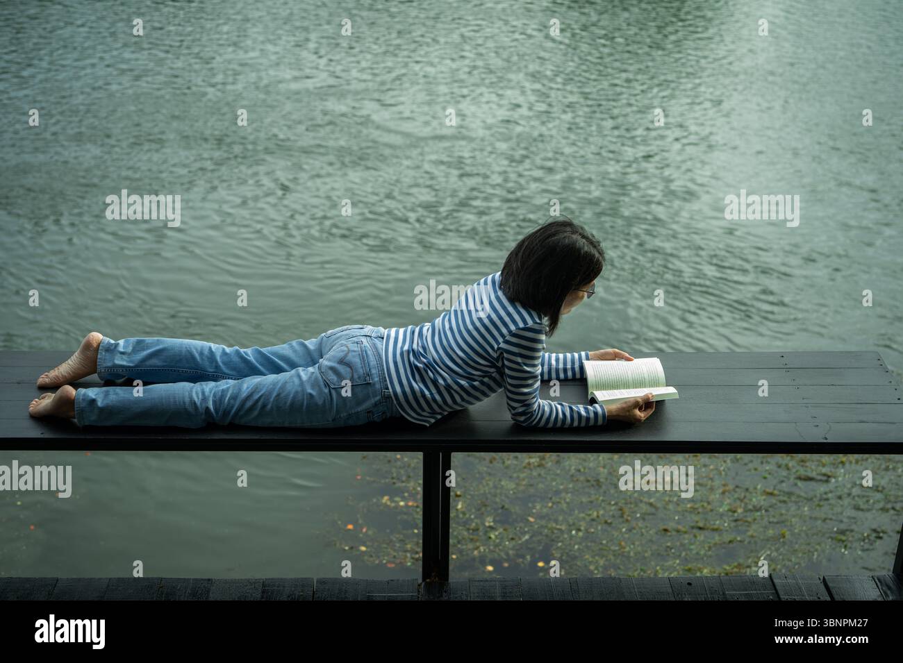 Eine Frau, die auf einer Holzterrasse am See liegt und ein Buch liest. Langsames Leben und digitale Entgiftung in der Natur ziehen sich für Selbstpflege und Balance zurück. Nachhaltiges selbst Stockfoto