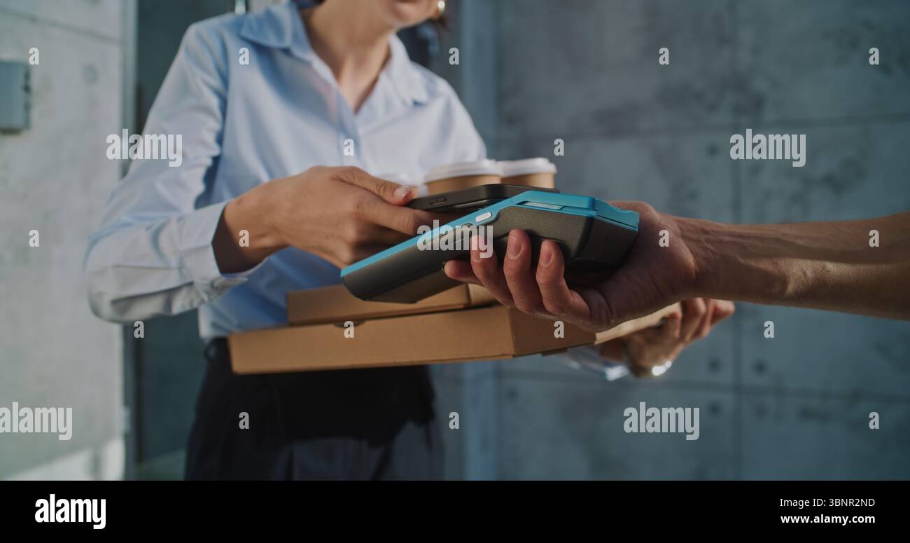 Liefermann übergibt Geschäftsfrau Zahlungsterminal nach erfolgreicher Pizzalieferung zum Bürogebäude. Frau hält Essen aus dem Restaurant, bezahlt für Bestellung kontaktlos mit Smartphone. Nahaufnahme. Stockfoto