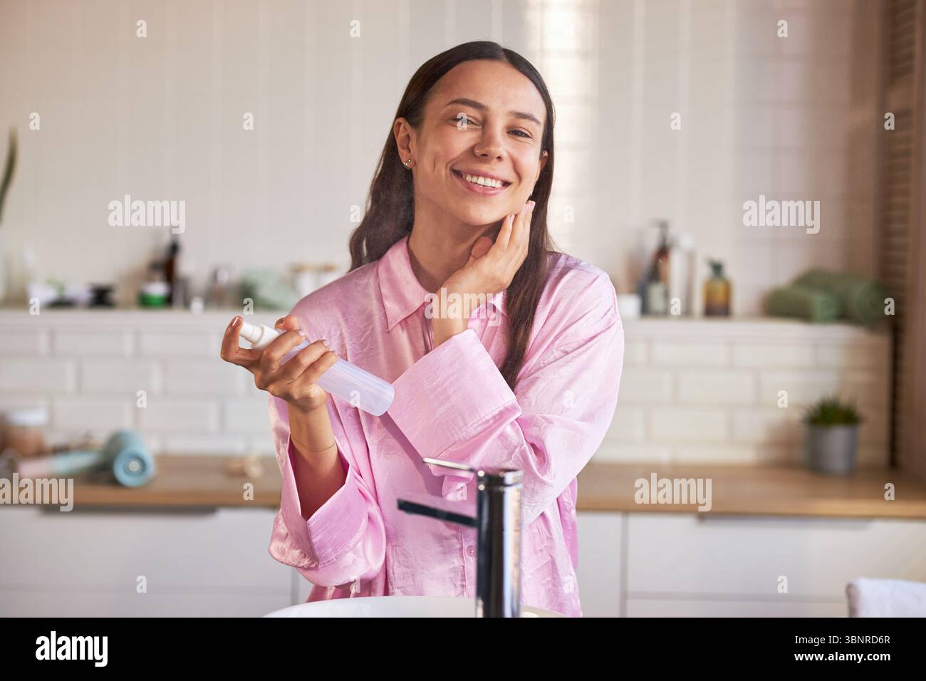 Porträt einer jungen Erwachsenen kaukasischen Frau lächelt beim Auftragen von Gesichtsnebel und Berühren der Wange während der morgendlichen Hautpflege-Routine im Badezimmer und demonstriert gesunde Schönheitsgewohnheiten Stockfoto