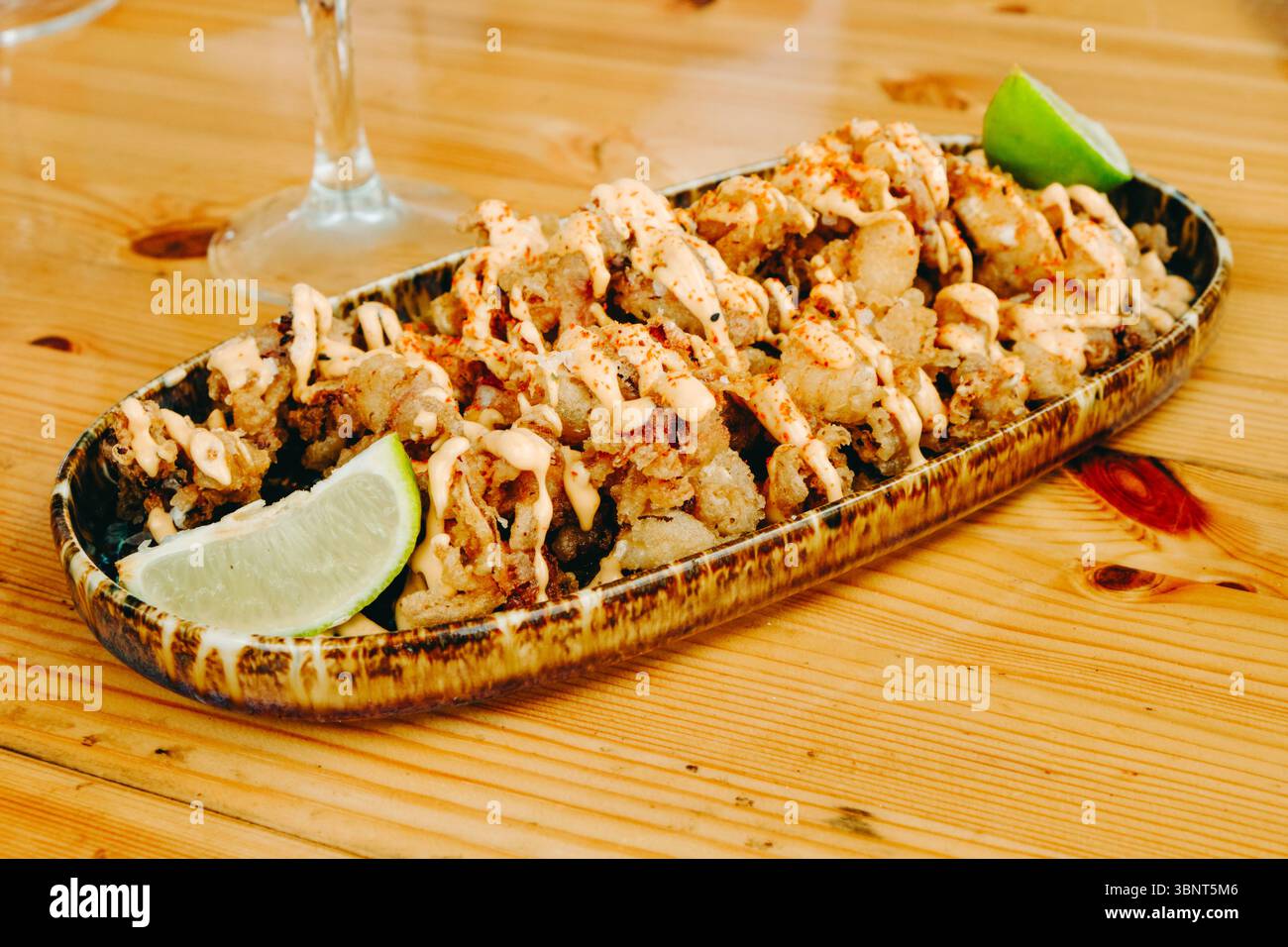 spanische Chipirones a la andaluza, knuspriger gebratener Baby-Tintenfisch mit Limettenkeilen und einem Spritzer Soße, serviert auf einem rustikalen Holztisch in einem hellen Innenraum Stockfoto