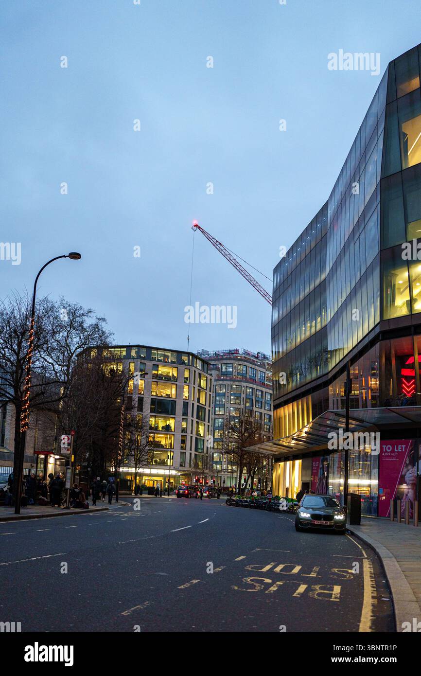 Vereinigtes Königreich, London, 11. Dezember 2024. Der abendliche Verkehr fließt durch ein modernes Geschäftsviertel mit Büro- und Verkaufsflächen, die bei Dämmerung beleuchtet sind. Stockfoto