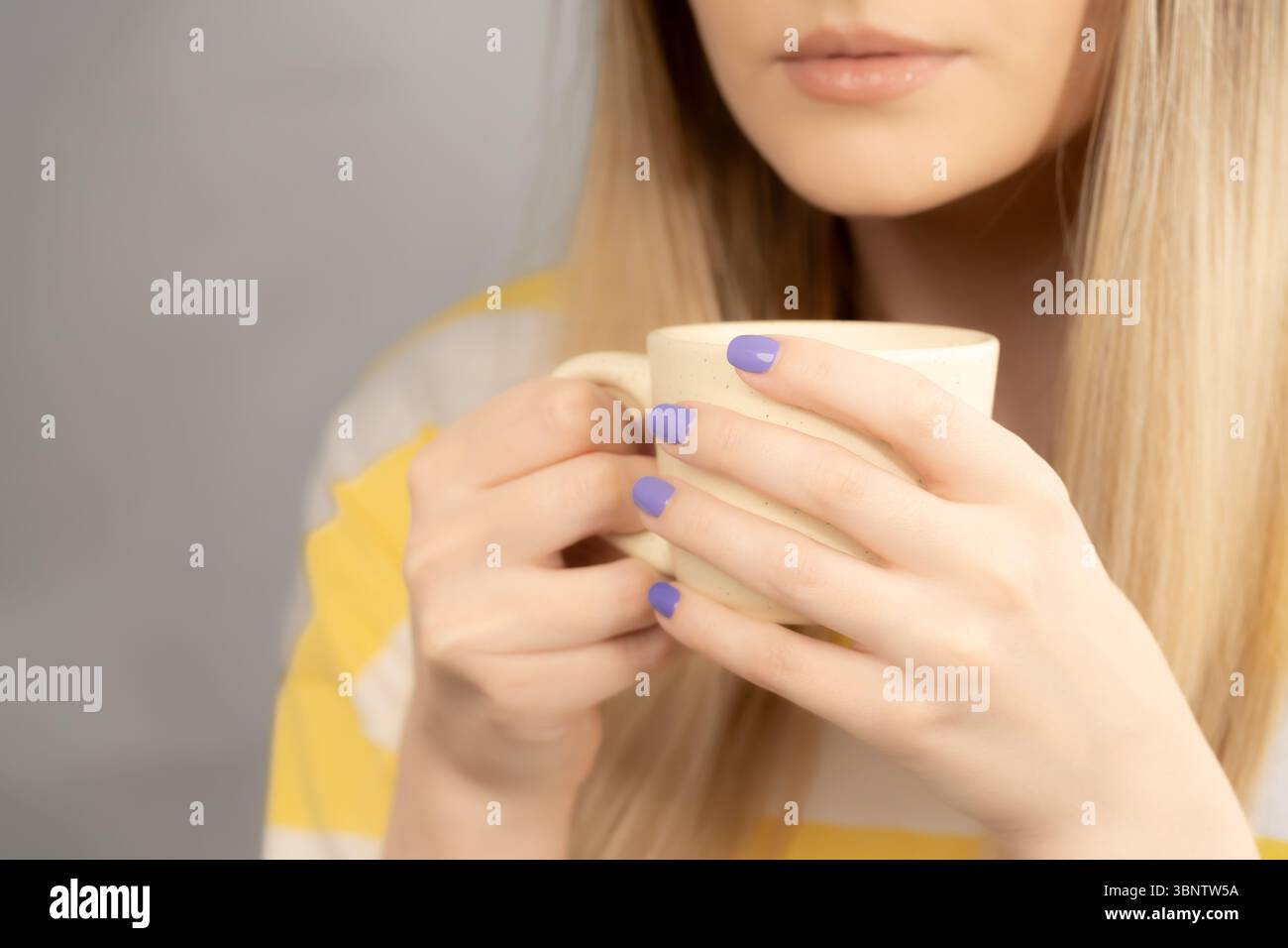 Eine Frau in einem gelb-weißen T-Shirt trinkt Kaffee mit Milch aus einer blassgelben Tasse. Der untere Teil ihres Gesichts ist in einer extremen Nahaufnahme zu sehen. Stockfoto